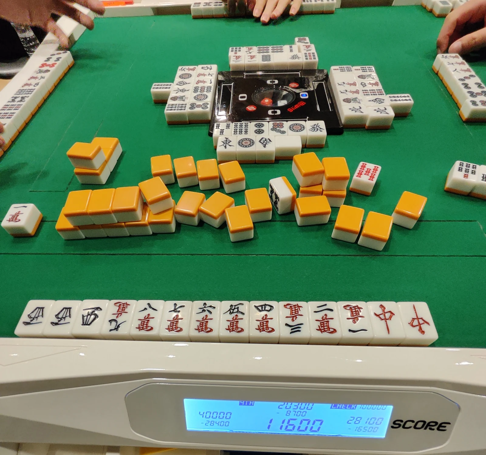 A riichi table mid-hand, with a large discard pile of face-down tiles, the four players' hands laid out, and the automatic table's score display in front