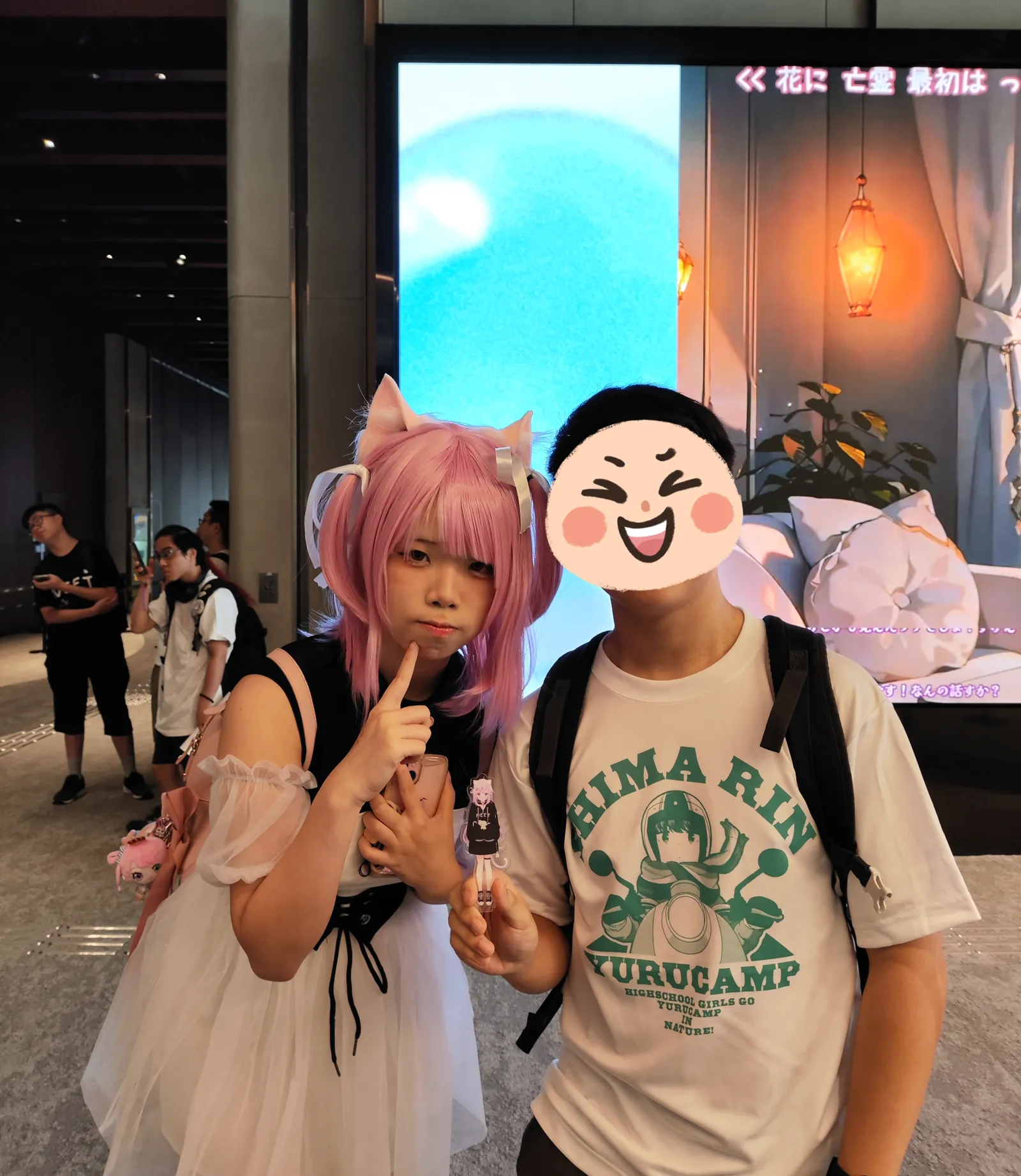 A Mikeneko cosplayer in a pink wig and white dress holding a small figurine, posing for a photo in front of the venue's screen